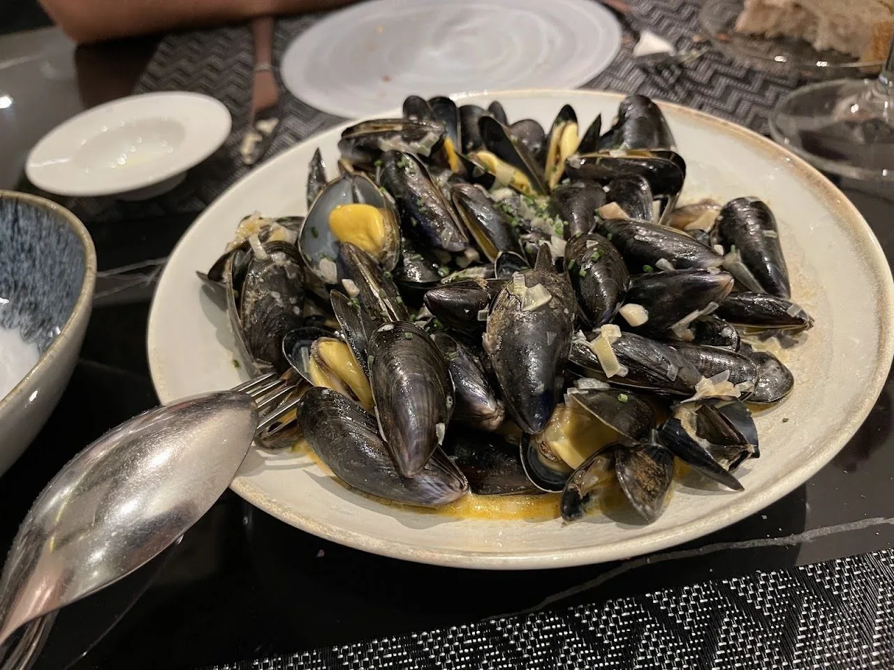 A white plate of open black mussels in a pale sauce with a serving spoon