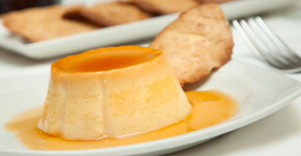 Flan de-molded on a white plate with caramel sauce pooling around it, a fried tuile cookie beside it