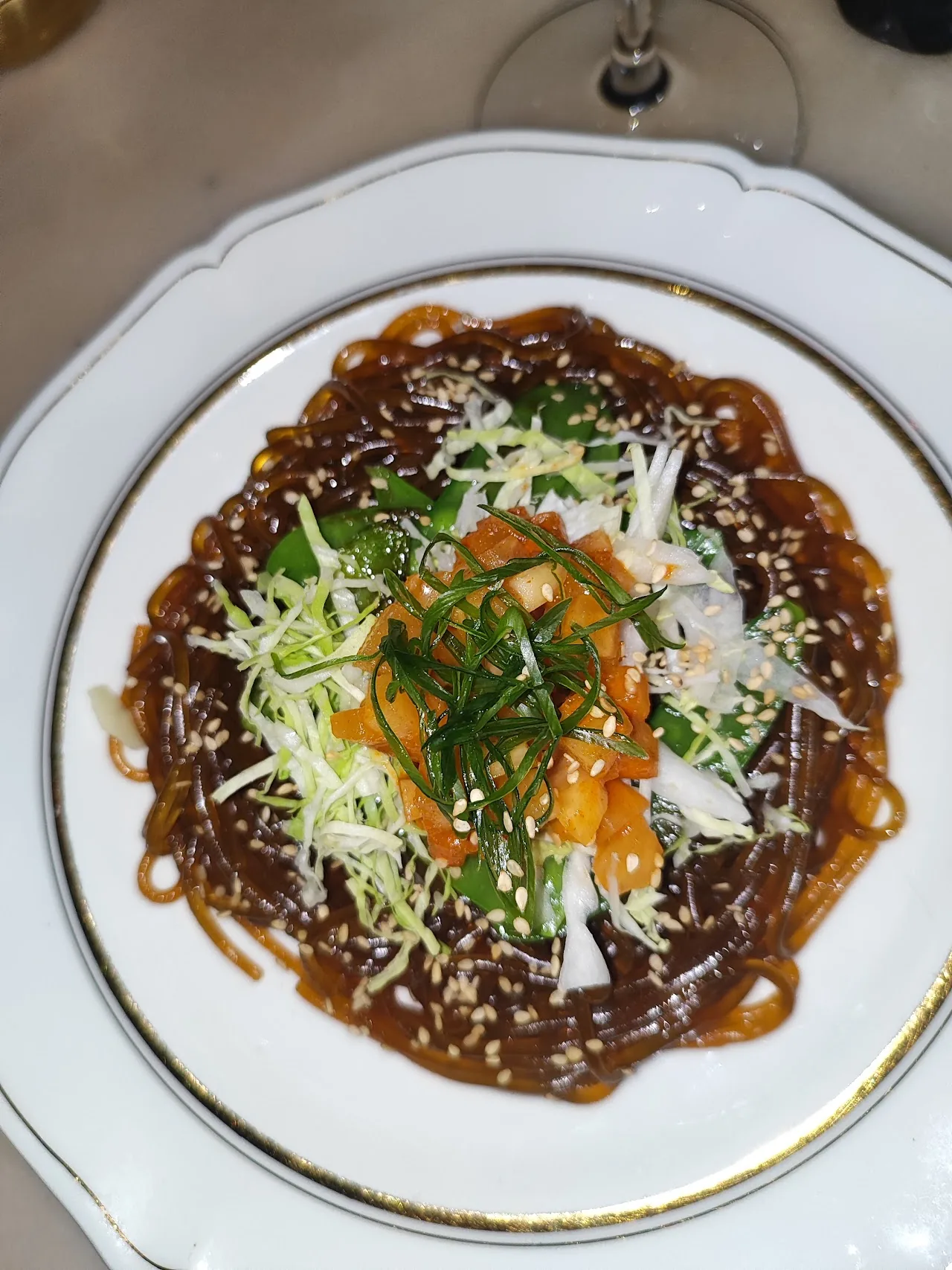 A plate of glossy dark-sauced noodles topped with shredded cabbage, snap peas, and diced kimchi, with a wine glass in the background