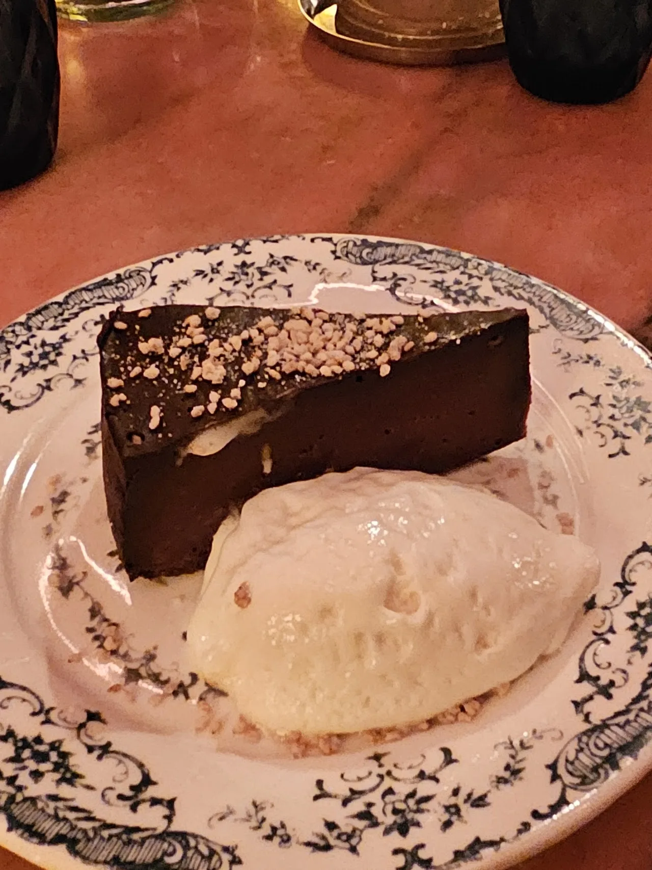 A wedge of dark chocolate cake topped with crushed nuts, served beside a scoop of cream on a floral-patterned plate