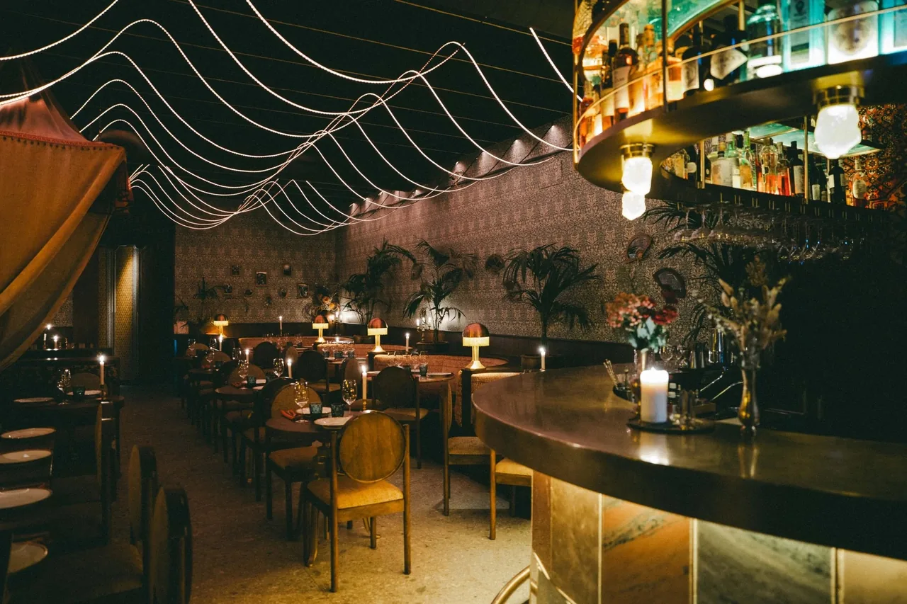 A dark dining room with candles on the tables, string lights on the ceiling and a round lit bar