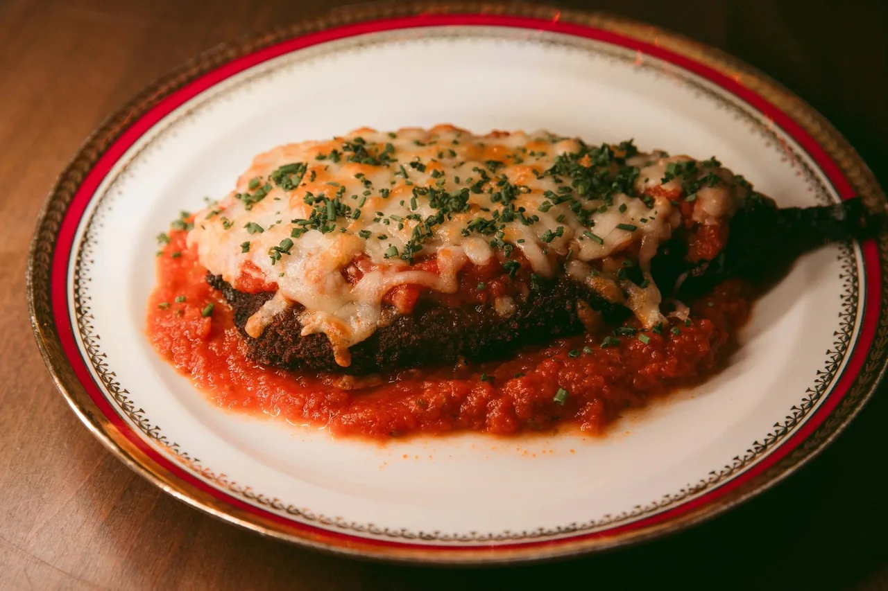 A gold-rimmed white plate with a baked aubergine dish in tomato sauce on a marble table