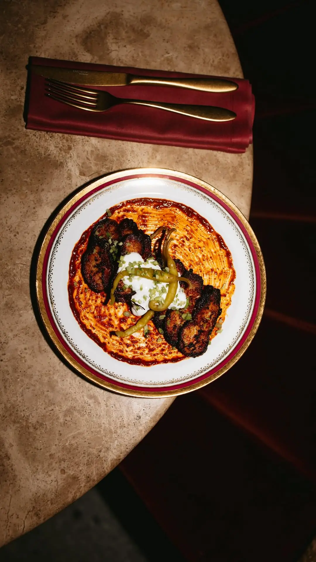 A plate of seared meat over red sauce with yogurt, pickled peppers, and scallion, with cutlery on a napkin