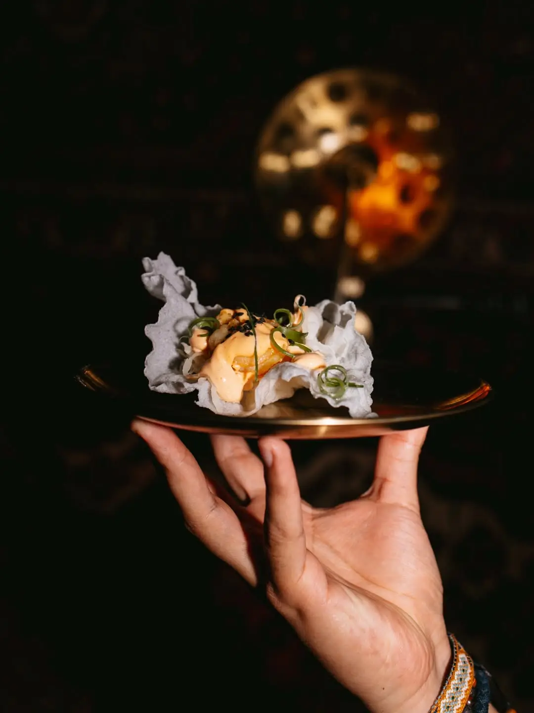 A hand holding a small plate with a crispy chip topped with sauce, herbs, and sesame seeds